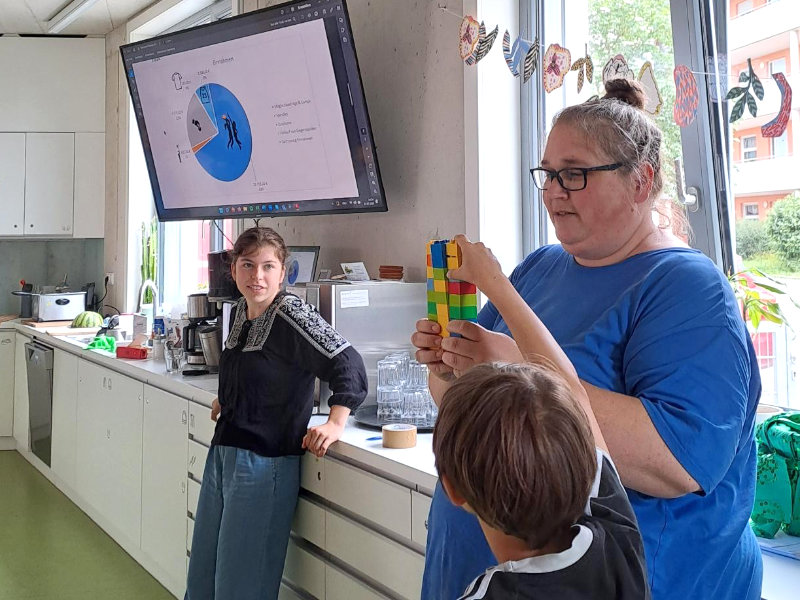 Zwei Vorstandsmitglieder von Artio zeigen anschaulich, wie die Einnahmen sich zusammen setzen: dafür ergänzt ein Kind einen Steckklotz auf einer Reihe von Steckklötzen.