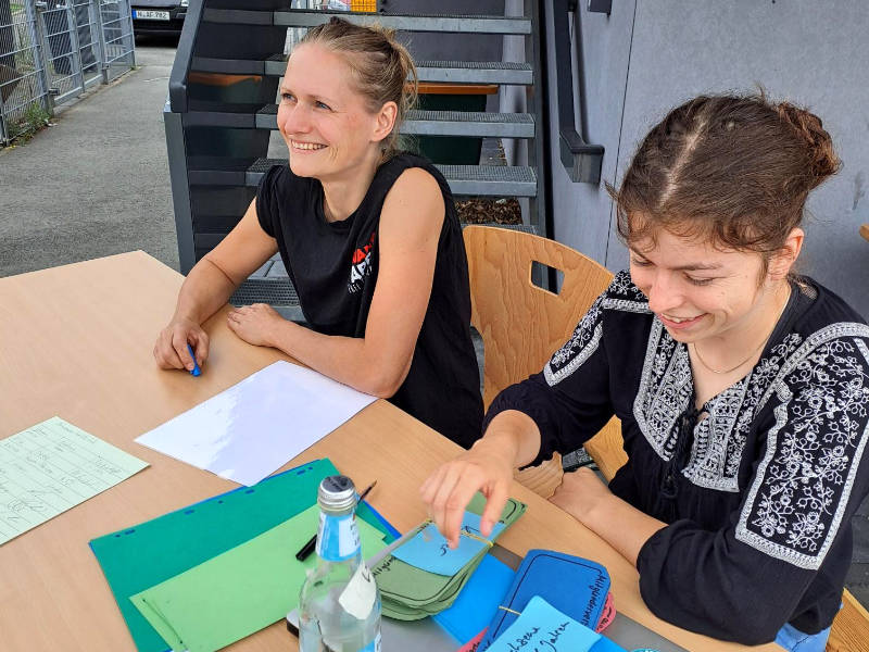Jasi und Lena vom Vorstand begrüßen alle Teilnehmenden: alle Neuankömmlinge registrieren sich in der Anwesenheitsliste und bekommen als Mitglieder ihre Stimmkarten ausgehändigt.