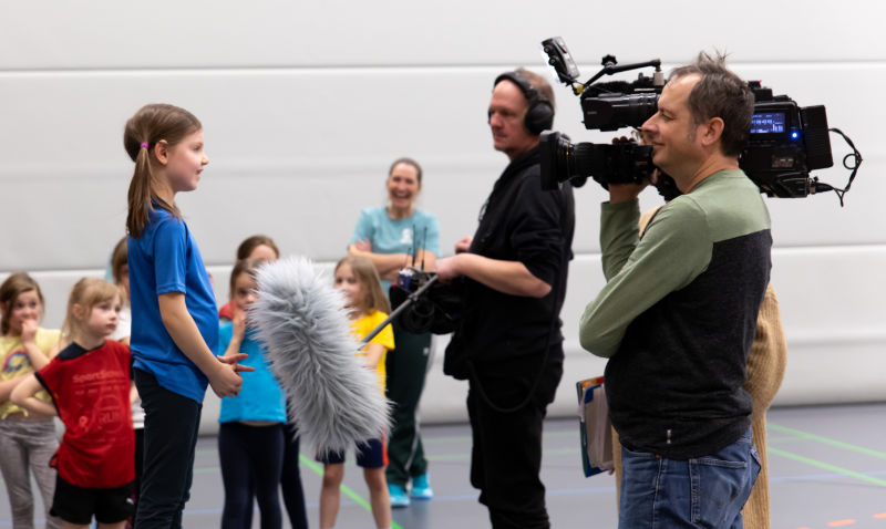 Ein Mädchen der U8 gibt ein Interview vor der Kamera. Im Hintergrund steht ihr Team und schaut gebannt zu.